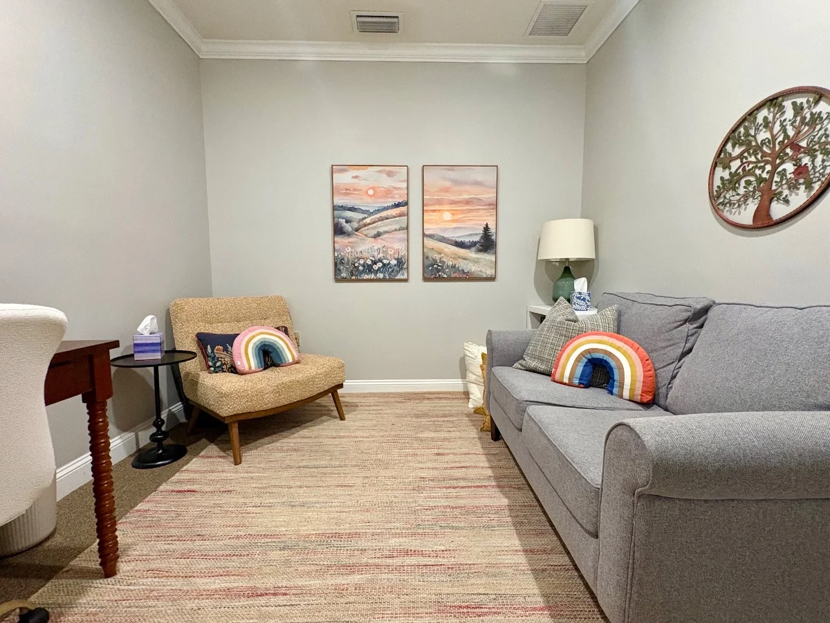 Therapy room with a grey sofa, a boucle armchair with a rainbow pillow, and two sunset landscape paintings